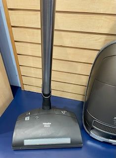 A Certified Refurbished Sew & Vac Miele C3 Brilliant vacuum cleaner head and handle leaning against a wooden slat wall next to a gray electronic device on a blue carpeted floor.