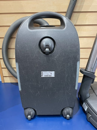 A certified refurbished Miele C3 Brilliant grey, upright vacuum cleaner with a hose attached, displayed in front of a wooden wall.