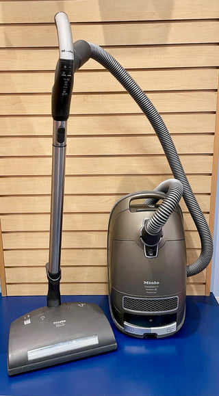 A Certified Refurbished Sew & Vac Miele C3 Brilliant vacuum cleaner with a hose and floor attachment, positioned against a wooden slat background.