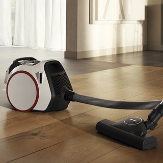 The 2-Miele Boost CX1 Pure Suction bagless canister vacuum cleaner with Vortex technology, equipped with a hose and floor brush attachment, is placed on a wooden floor in a brightly lit room.