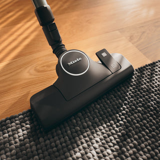The 2-Miele Boost CX1 Pure Suction vacuum cleaner head is shown on a textured gray rug and hardwood floor under natural light, highlighting its advanced Vortex technology and highly effective AirClean filter for optimal dust capture.