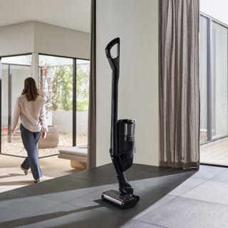 The Miele Triflex HX3 Cordless Stick Vacuum in Brilliant White stands upright on a dark tile floor in a modern room as a person walks away toward large windows and curtains.