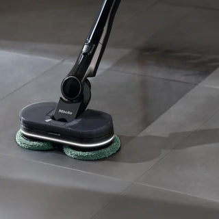 A floor cleaning machine, the Miele Triflex HX3 Aqua Cordless Wet & Dry Vacuum in Obsidian Black by 2-Miele, is shown cleaning a tiled floor and leaving a visible clean path behind it.