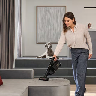 A woman uses the 2-Miele Miele Triflex HX3 Cat & Dog Cordless Stick Vacuum in Obsidian Black to effortlessly clean her couch, while her dog relaxes on a raised platform in the background.