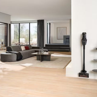 Modern living room with large windows, a sectional sofa, coffee table, and a Miele Triflex HX3 Aqua Plus Cordless Wet & Dry Vacuum in Obsidian Black by 2-Miele mounted on the wall near shelving, featuring a hard-floor roller for quick and efficient cleaning.