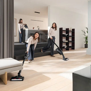 Three women, dressed similarly, use the 2-Miele Miele Triflex HX3 Cordless Stick Vacuum in Brilliant White to clean a modern living room and kitchen area, highlighting its versatility for simultaneously tackling multiple cleaning tasks.