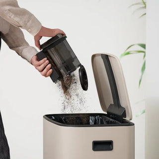 A person empties coffee grounds from a French press into a beige trash bin with a black interior, preparing to clean up any remaining mess with the Miele Triflex HX3 Cordless Stick Vacuum in Brilliant White by 2-Miele.