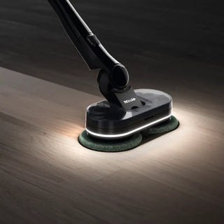 Close-up of a 2-Miele Triflex HX3 Aqua Cordless Wet & Dry Vacuum in Obsidian Black with two rotating pads and LED lights, shown cleaning a dark wooden floor.