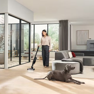 A woman uses the 2-Miele Miele Triflex HX3 Cat & Dog Cordless Stick Vacuum in Obsidian Black to clean a bright, modern living room with large windows, while a gray dog relaxes on the floor in the foreground.