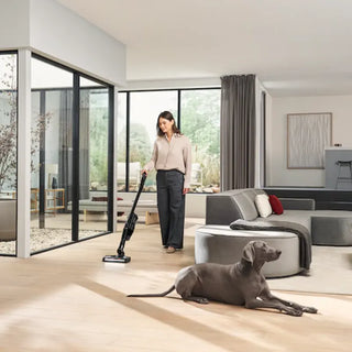 A woman vacuums the floor in a modern living room with large windows using the Miele Triflex HX3 Cordless Stick Vacuum in Brilliant White by Miele, while a gray dog lies on the light wood floor nearby.