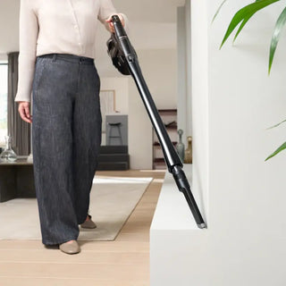 A person vacuums a white ledge in a modern living room using the 2-Miele Miele Triflex HX3 Cordless Stick Vacuum in Brilliant White, featuring a HEPA filter.