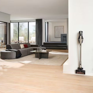 The Miele Triflex HX3 Plus Cordless Stick Vacuum in Titanium Pearl by 2-Miele, known for its long battery life, is mounted on a white wall in a modern living room featuring large windows, a gray sofa, coffee table, and light wood flooring.