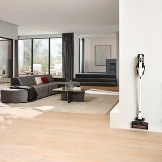 The Miele Triflex HX3 Cordless Stick Vacuum in Brilliant White by Miele, featuring a HEPA filter, stands upright against a white wall in a modern living room with large windows, a gray sofa, and neutral decor.