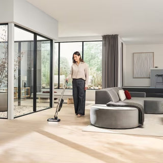 A woman vacuums a light wood floor in a modern, bright living room with large windows, gray furniture, and minimalist decor, using the Miele Triflex HX3 Plus Cordless Stick Vacuum - Titanium Pearl by 2-Miele, which is equipped with a HEPA filter.