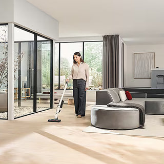 In a modern, bright living room with large windows and minimalist furniture, a woman cleans the floor using the Miele Triflex HX3 Cordless Stick Vacuum in Brilliant White, featuring advanced FloorDetect technology.