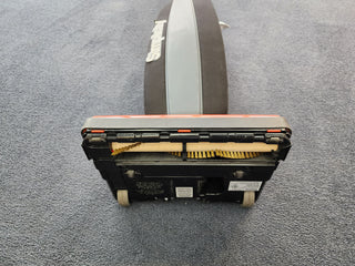 A surfboard resting on a carpeted floor while a Used Simplicity S10CV Cordless Vacuum by 10-Tacony with a HEPA media bag nearby aids in charcoal filtration.