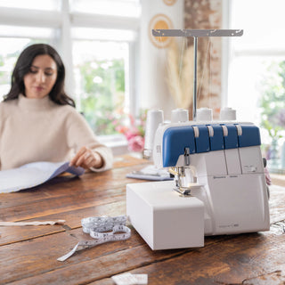 A woman works with fabric at a wooden table near a Husqvarna Viking Amber Air S400 Overlock Machine and a measuring tape.