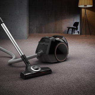 A modern bagless vacuum cleaner, the 2-Miele Boost CX1 Cat & Dog, sits on a carpeted floor in a dimly lit room, with a chair and lamp in the background. Designed for powerful suction and effective pet hair removal, this sleek model keeps your home spotless.