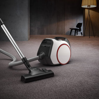 The 2-Miele Boost CX1 Pure Suction bagless canister vacuum, equipped with advanced Vortex technology and a hose with floor head, is positioned on a carpeted floor in a modern, dimly lit room featuring a chair and lamp in the background.