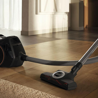 The 2-Miele Boost CX1 Cat & Dog bagless vacuum cleaner, featuring a long hose and brush head, is shown cleaning pet hair from a hardwood floor in a well-lit room with light-colored curtains, making pet hair removal effortless.