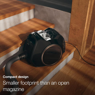 A compact 2-Miele Boost CX1 Cat & Dog vacuum cleaner rests on wooden stairs, its hose and cord extended in the sunlight. Text reads: “Compact design. Smaller footprint than an open magazine.” Ideal for pet hair, the Miele Boost CX1 Cat & Dog keeps your stairs spotless every time.