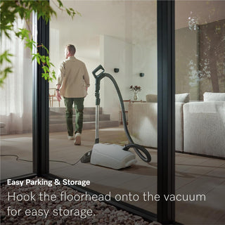 A person walks away from a 2-Miele Guard L1 Electro canister vacuum with the floorhead securely hooked onto the machine in a bright living room. Text reads: “Easy Parking & Storage. Simply hook the floorhead onto your Miele Guard L1 Electro for convenient storage.”.