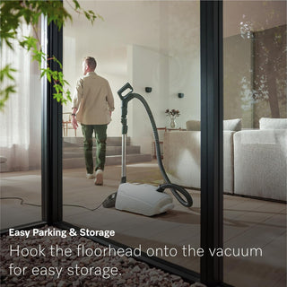A person walks away from a 2-Miele Guard L1 AllFloor bagged vacuum cleaner with its floorhead securely attached to the main unit in a contemporary living room. On-screen text showcases the hassle-free storage solution and emphasizes the convenience of AirClean Plus filtration, ensuring a healthier home environment.