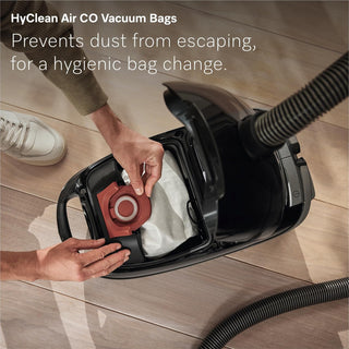 A person inserts a red vacuum bag into the sleek black 2-Miele Guard M1 Cat & Dog bagged canister vacuum on a wood floor, with text highlighting hygienic, dust-free bag changes and the added benefit of an Active AirClean filter for superior pet-hair cleaning.