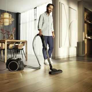 A man uses the 2-Miele Boost CX1 Cat & Dog bagless vacuum cleaner on a wooden floor in a bright, modern dining and living room, effortlessly and efficiently removing pet hair.