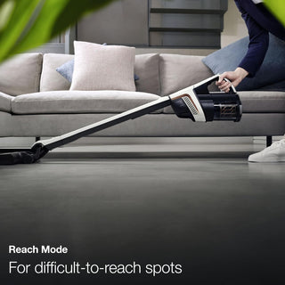 A person uses the Miele TriFlex Lotus White by Sew & Vac, featuring an extended handle and a Digital Efficiency Motor, to clean under a couch. Text reads: "Reach Mode: For difficult-to-reach spots and powerful cleaning.