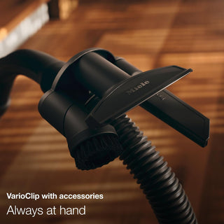 Close-up of a 2-Miele Boost CX1 Cat & Dog bagless vacuum cleaner with the VarioClip accessory holder attached to the hose, displaying the brush and crevice tool—perfect for swift pet hair cleanup. Text reads: “VarioClip with accessories. Always at hand.”.