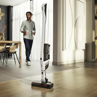 A man holding two mugs walks through a modern living space with wooden floors, while a sleek, white Miele TriFlex Lotus White vacuum cleaner by Sew & Vac stands prominently in the foreground.