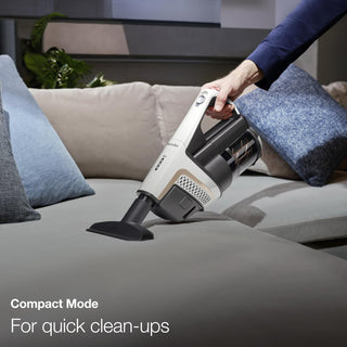 A person uses a Miele TriFlex Lotus White vacuum from Sew & Vac on a beige couch with blue and gray cushions. Text reads, "Compact Mode for quick clean-ups with Triflex 3in1 design.