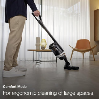 A person is effortlessly using the Sew & Vac Miele TriFlex Lotus White vacuum cleaner with a Digital Efficiency Motor on the living room floor, ensuring every corner beneath the chair and table is spotless.