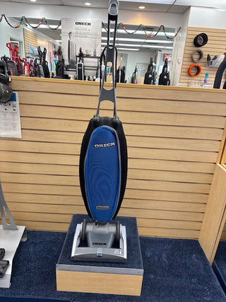 A blue Oreck Magnesium RS Swivel-Steering Upright Vacuum Cleaner, certified reconditioned, is showcased in a store on a blue carpeted surface. This lightweight vacuum captivates attention amidst a backdrop of shelves displaying similar models.