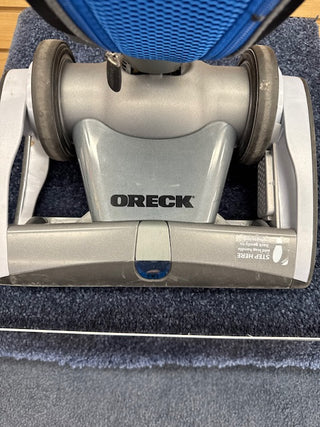 Close-up of the Oreck Magnesium RS Swivel-Steering Upright Vacuum RLW1500RS Certified Reconditioned head on a dark carpet, highlighting its SlimSwivel Design for effortless maneuverability.