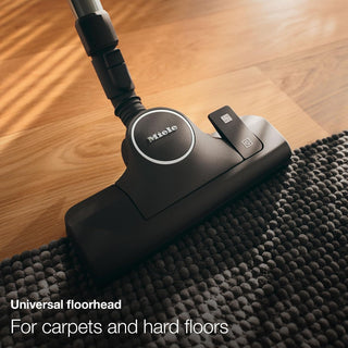 A 2-Miele Boost CX1 Cat & Dog bagless vacuum cleaner with a universal floorhead is displayed on the border between a textured rug and hardwood flooring, with text emphasizing its strong pet hair removal performance on both carpets and hard floors.