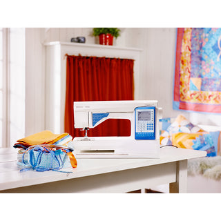 A Husqvarna Viking Sapphire 930 sewing machine by Husqvarna Viking is on a white table with colorful fabric, thread spools, and scissors in a bright room with a red curtain and a quilt hanging on the wall.