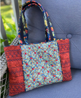 A **Bag of Tricks quilted tote bag** featuring intricate embroidery designs in red and blue, displayed on a chair.