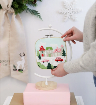 Someone holds an embroidery hoop with a festive winter village—houses, trees, red cars, and snowflakes—in the background. Ideal for fans of holiday designs from the Sew & Vac Kimberbell Comfort & Joy 2 Day Event.