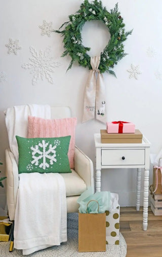 A cozy white armchair with blankets and snowflake pillows sits by a small table of wrapped gifts. Festive wreaths and snowflakes frame the scene, showcasing Sew & Vac’s Kimberbell Comfort & Joy- 2 Day Event holiday designs.