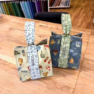 Two fabric tote bags made in the "THE GIVING BAG" Reusable Gift Bag Class with Carla (Layton In-Store Class, 12/6/25, 1–4 PM) are shown on a wooden table—perfect for beginner sewing projects or as eco-friendly gift bags.