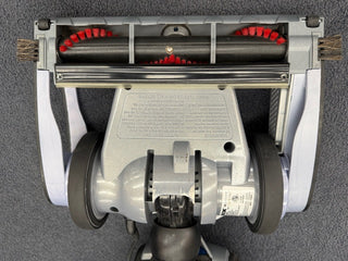 Bottom view of a Sew & Vac certified refurbished Oreck Magnesium LW1500RS vacuum head featuring two wheels, a red-bristle roller brush, side brushes, and manufacturer info on a gray background.