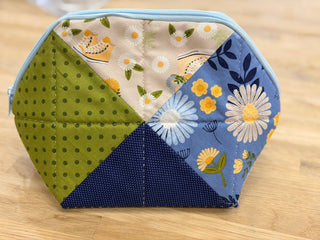 A Minki Kim Criss Cross Pouch, crafted from six triangular fabric panels in blue, green, beige, and floral prints, rests on a wooden surface—an inspiring project you can create during the Layton In-Store Class: Criss Cross Pouch Set with Minki Kim on Saturday, September 19th from 1–4 pm at Imagine Sewing & Vacuum’s Layton location.