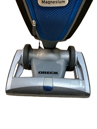 Close-up view of the base of a refurbished Oreck Magnesium vacuum by Imagine Sewing & Vacuum, showing the brand name and part of the blue HEPA bag.