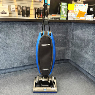 A 1-Oreck LW100 Magnesium Lightweight Vacuum (Certified Refurbished), featuring HEPA filtration and a blue/black design, is shown on a carpeted floor in an electronics store.