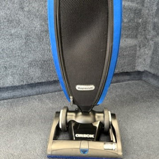 A 1-Oreck LW100 Magnesium Lightweight Vacuum (Certified Refurbished) in black and blue, labeled "Magnesium," is placed on a gray carpeted floor, highlighting its light design.
