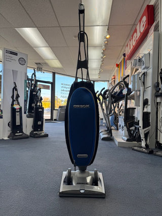 An Oreck Magnesium RS Swivel-Steering Upright Vacuum RLW1500RS Certified Reconditioned is displayed on the floor of an appliance store, with other vacuums visible in the background.