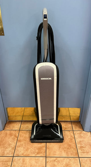 A Certified Refurbished Sew & Vac Oreck Elevate Conquer upright vacuum cleaner positioned on a tiled floor against a blue wall.