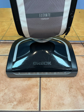 An Sew & Vac Oreck Elevate Conquer vacuum cleaner on a tiled floor with a grey and white suitcase behind it displaying the text "elevate conquer," featuring a Certified Refurbished HEPA CC bag.
